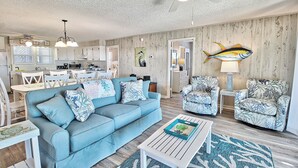 A spacious living area with a blue sofa, white coffee table, and patterned armchairs. A kitchen is visible in the background with white cabinets and a dining area with white chairs. - Oceanfront Ground Level Duplex, Beach Boardwalk, Screened Porch, Dog Friendly (Pawleys Island)