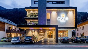 A modern hotel with a prominent entrance, multiple parking spaces, and a view of the surrounding forest. - Post Hotel (See)