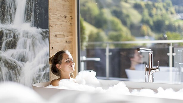 A woman in a bathtub filled with bubbles, with a waterfall and a view of greenery outside. - Post Hotel (See)