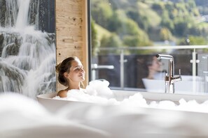 A woman in a bathtub filled with bubbles, with a waterfall and a view of greenery outside.