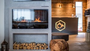 A modern interior with a fireplace, wooden logs, a stone accent piece, and a reception desk with a logo. - Post Hotel (See)
