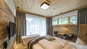 A modern hotel room with a wooden ceiling, a large window with curtains, a bathtub, a bed with pillows, a desk with chairs, and a TV mounted on the wall. - Post Hotel (See)