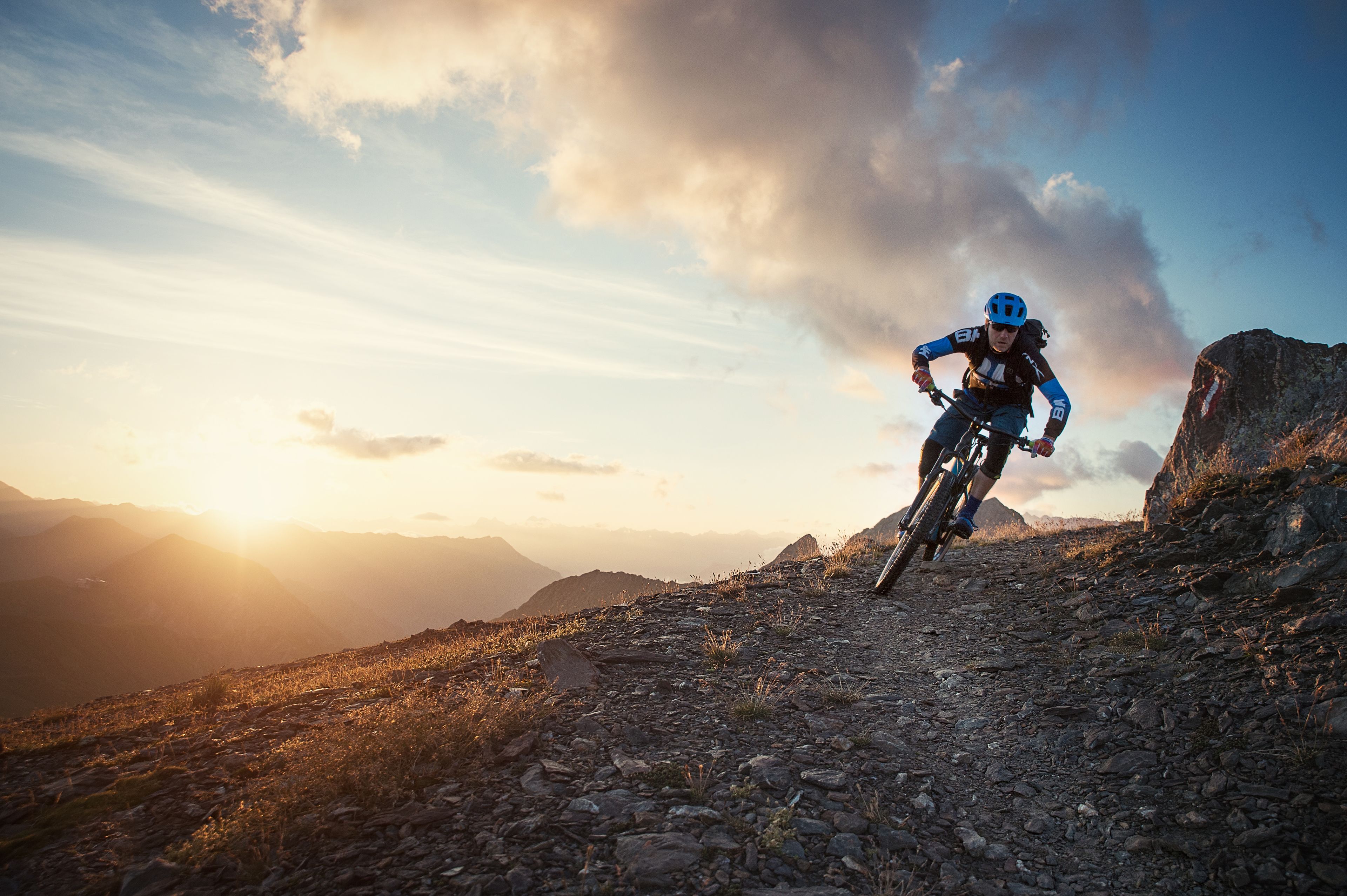 A mountain biker on a rocky trail with a mountainous backdrop and a sunset.
