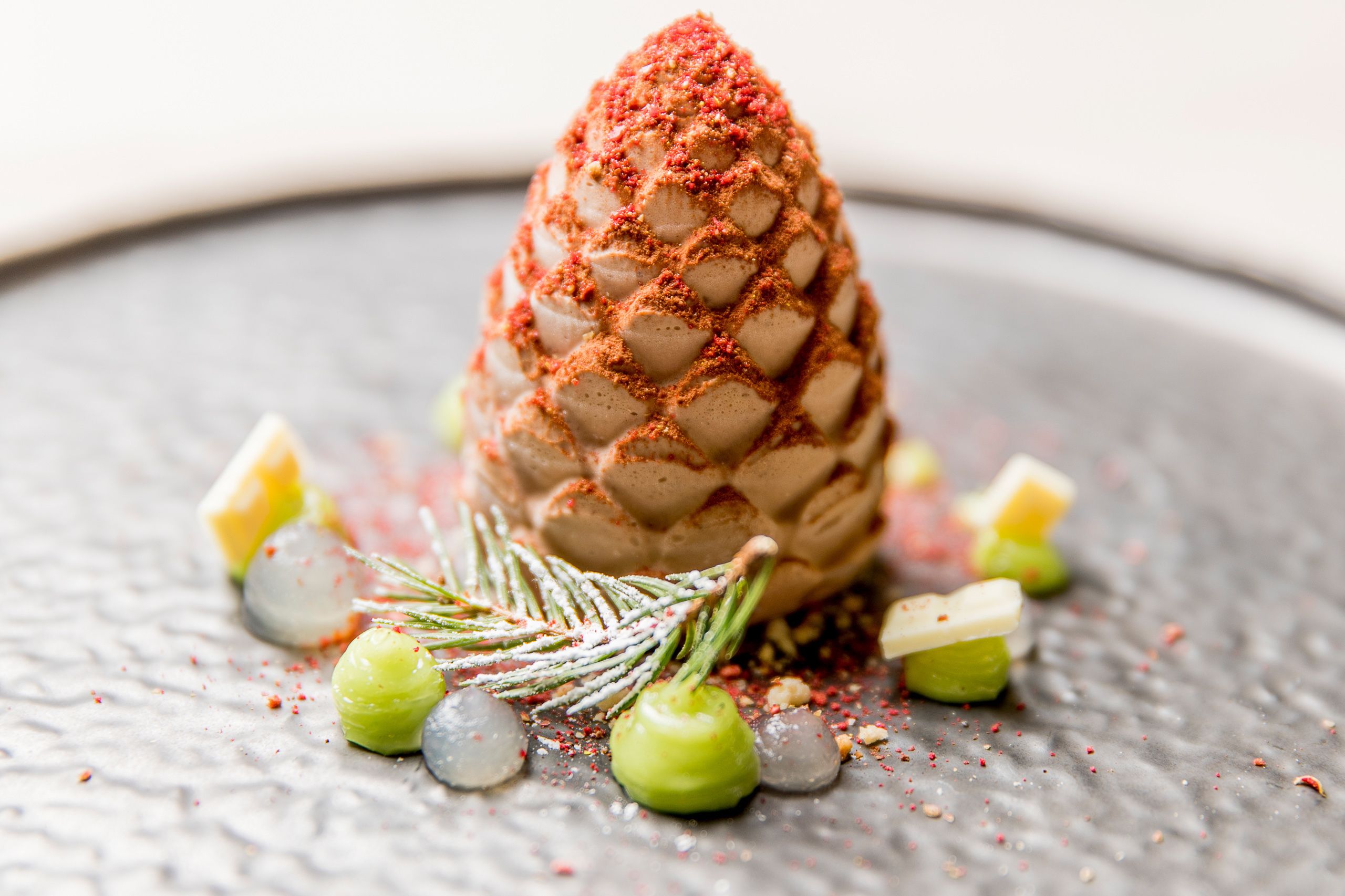 A dessert with a textured cone shape, garnished with green and white elements and sprinkled with red powder.