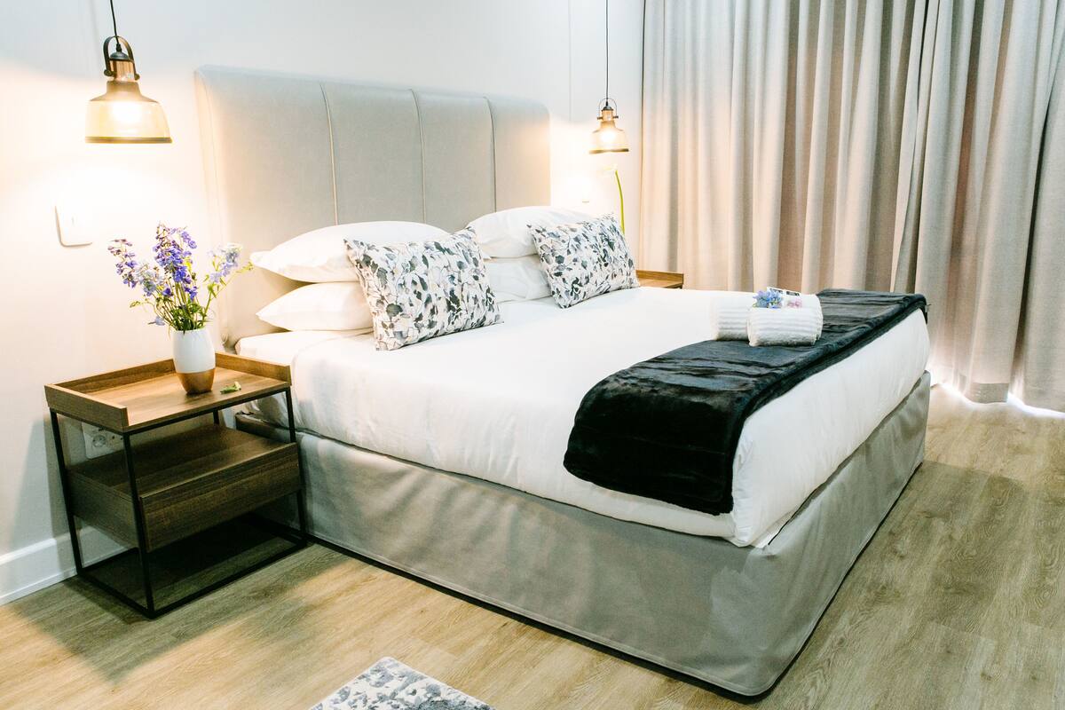 Una habitación de hotel moderna con una cama grande, una mesita de noche con un jarrón de flores y dos lámparas colgantes.