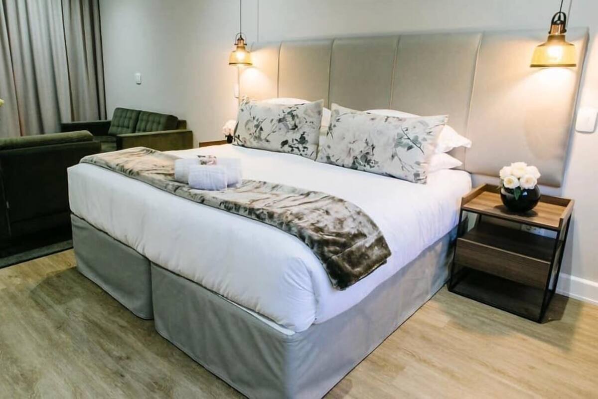 Una habitación de hotel moderna con una cama grande, dos lámparas de noche, una mesita de noche de madera y un sillón verde.