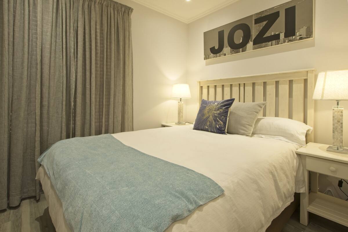 A neatly made bed with a blue throw blanket, two bedside lamps, and a pillow with a blue and gold design. A large sign with the word "JOZI" is on the wall above the bed.
