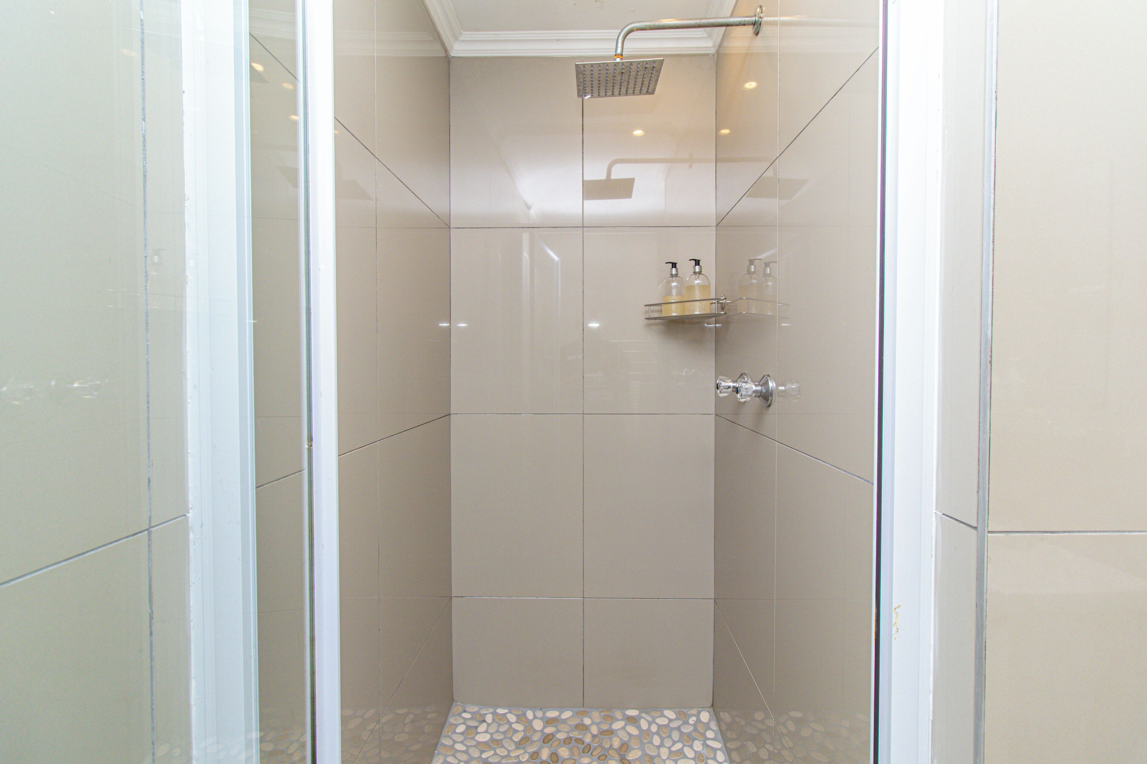 A glass-enclosed shower with a handheld showerhead, a glass shelf with toiletries, and a pebble-patterned floor.