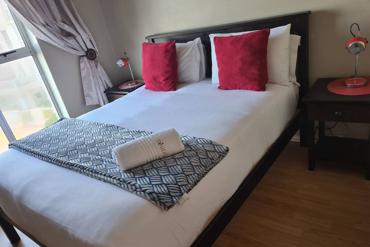 A neatly made bed with a patterned throw, two red pillows, and a white towel.