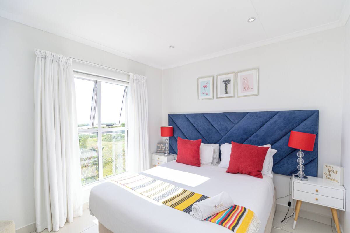 A hotel room with a large bed, white curtains, a blue headboard, and a window with a view.