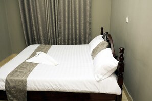 A neatly made bed with white linens and a gray accent, a wooden headboard, and a gray curtain partially drawn to the side.