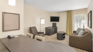 A hotel room with a large desk, two chairs, a sofa, a flat-screen TV, a small table, and a window with curtains.