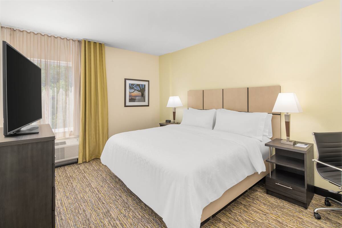 Une chambre d’hôtel avec un grand lit, deux tables de chevet avec des lampes, un téléviseur posé sur une commode et une fenêtre avec des rideaux.