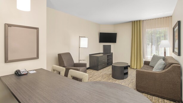A hotel room with a desk, two chairs, a sofa, a television, a flat-screen TV mounted on the wall, a telephone, and a lamp. - Candlewood Suites Newnan - Atlanta SW by IHG (Newnan)