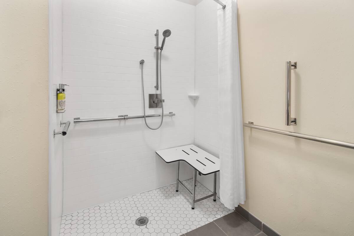 Une douche accessible aux personnes en fauteuil roulant, équipée d’une barre d’appui, d’un sol carrelé de couleur blanche et d’un petit banc.