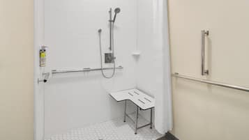 Un box de ducha accesible en silla de ruedas, con barra de apoyo, suelo de baldosas blancas y un pequeño banco.