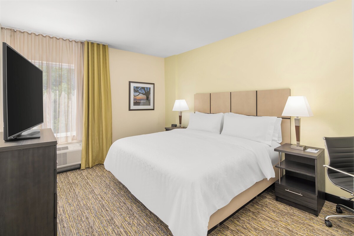 A hotel room with a bed, two bedside tables, a TV, a chair, and a window with curtains.