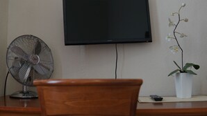 A room with a television mounted on the wall, a standing fan, a potted orchid, and a remote control on a wooden desk. - Hotel-Events Adlerpalast (Ruesselsheim)
