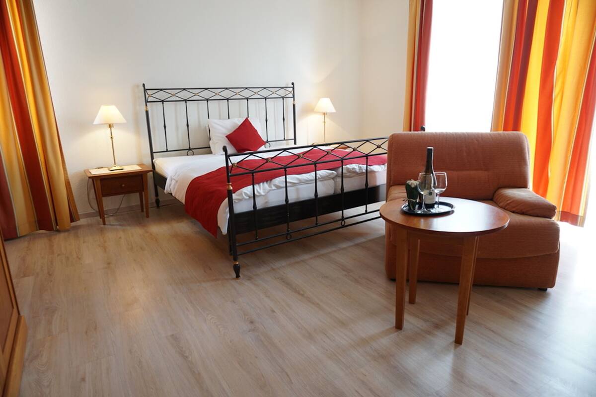 Une chambre d’hôtel avec un lit, des tables de chevet, une chaise et une petite table sur laquelle se trouvent une bouteille et un verre.