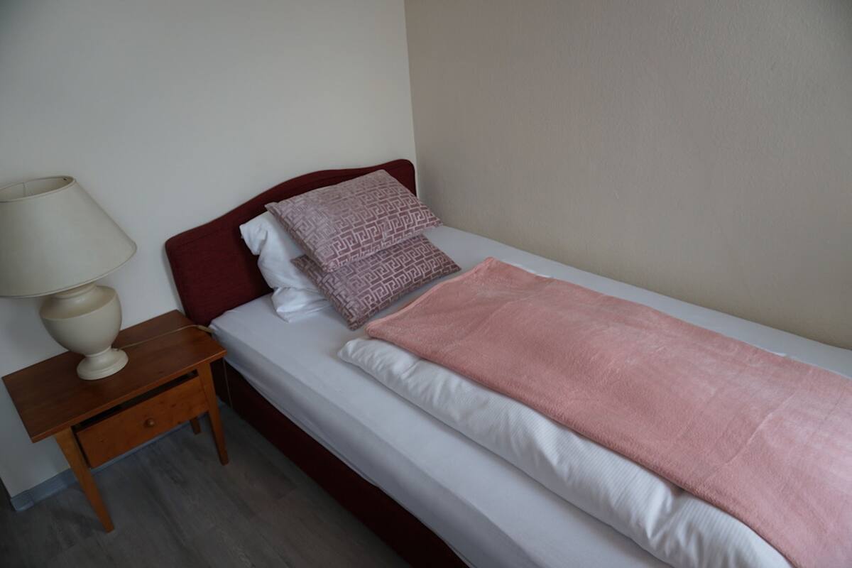 Un lit simple avec une tête de lit rouge, du linge de lit blanc et une couverture rose. Une table de chevet en bois avec une lampe se trouve à côté du lit.