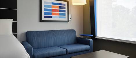 Ein Hotelzimmer mit blauer Couch, einem kleinen Tisch, einem gerahmten abstrakten Kunstwerk, einer Stehlampe und einem Fenster mit Blick auf Bäume.