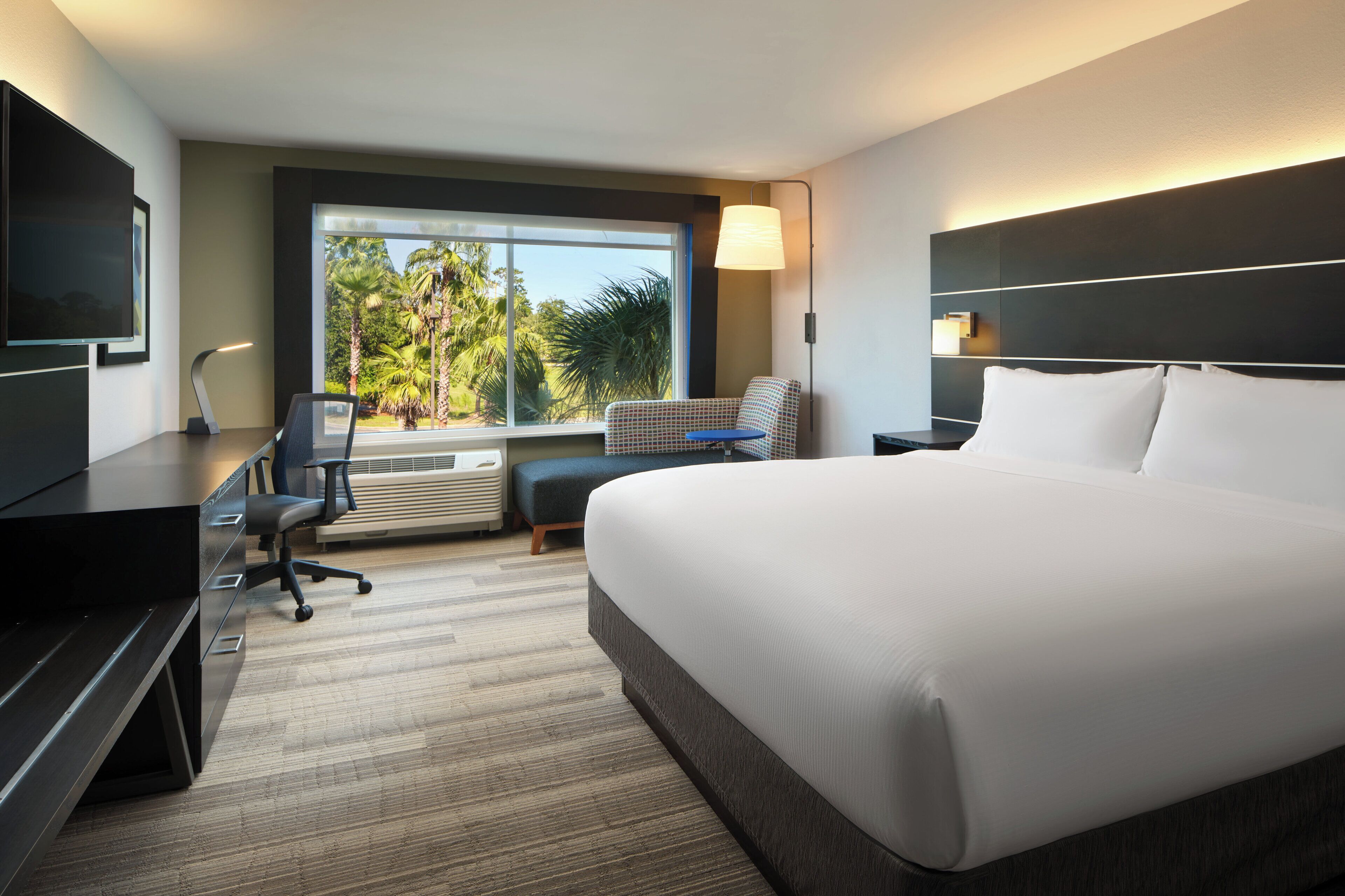 A modern hotel room with a large bed, a desk with a chair, a flat-screen TV, and a window with a view of palm trees.