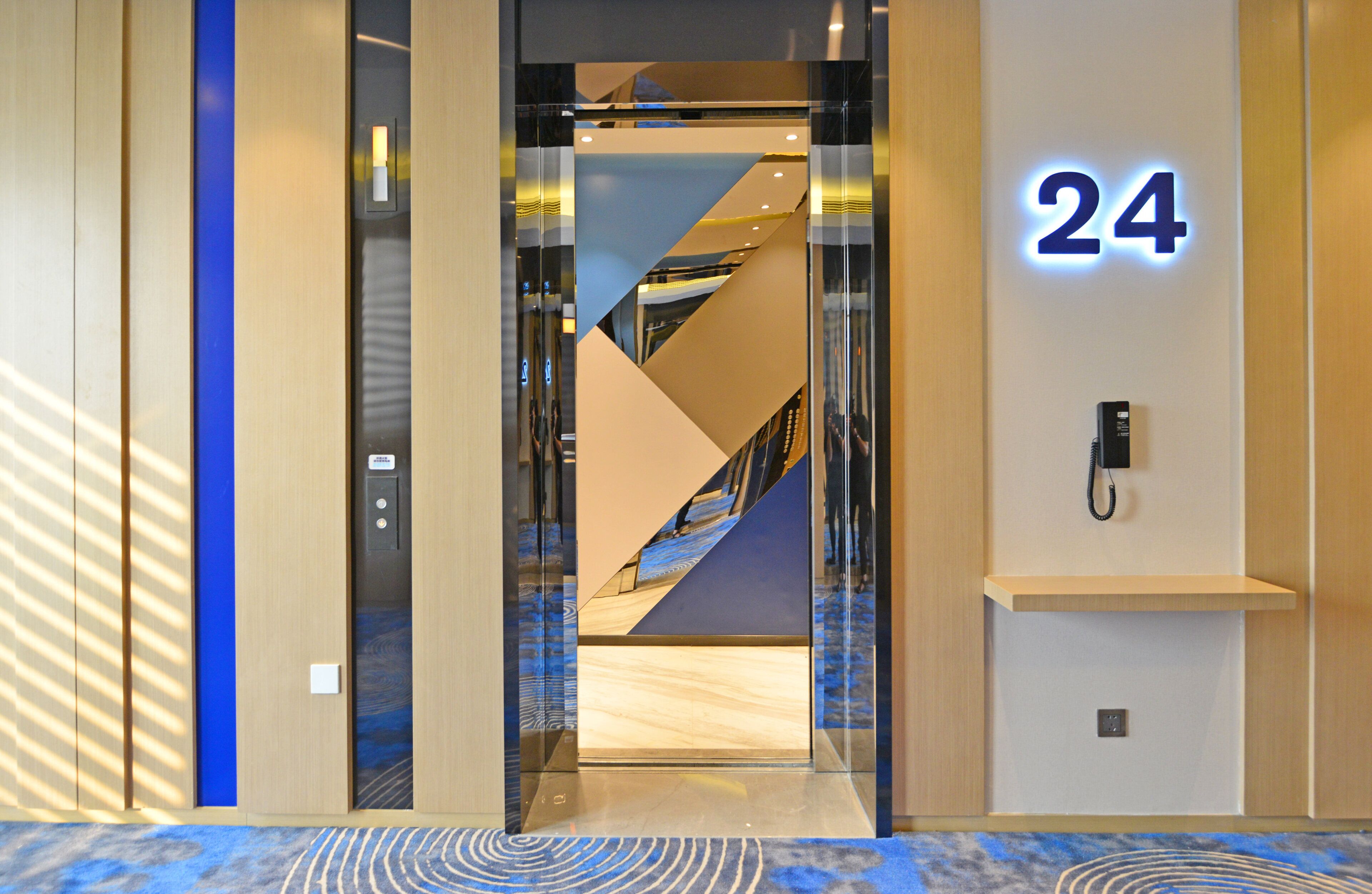 Vista interior de la entrada de un hotel con diseño moderno, que incluye un ascensor de vidrio, un letrero con el número '24' y un teléfono en la pared.