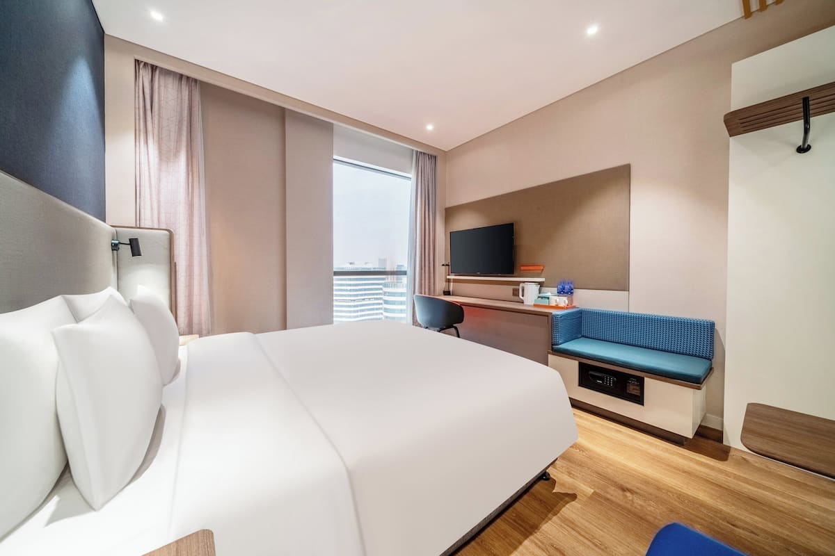 A modern hotel room with a large bed, a desk with a flat-screen TV, a seating area, and a view of the city.