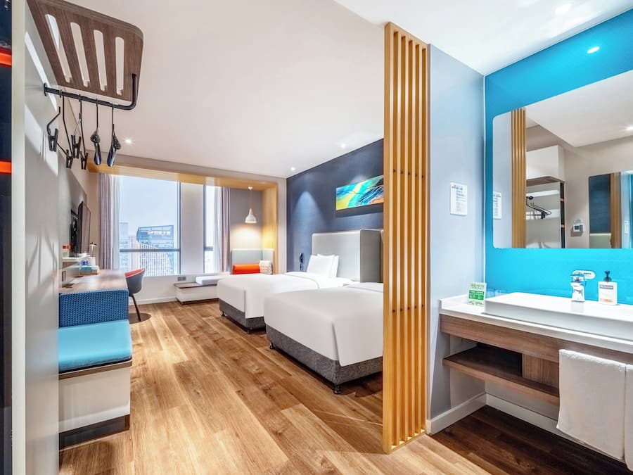 Habitación de hotel moderna con una cama grande, un escritorio, una silla, un baño con lavabo y espejo, y vistas a la ciudad.
