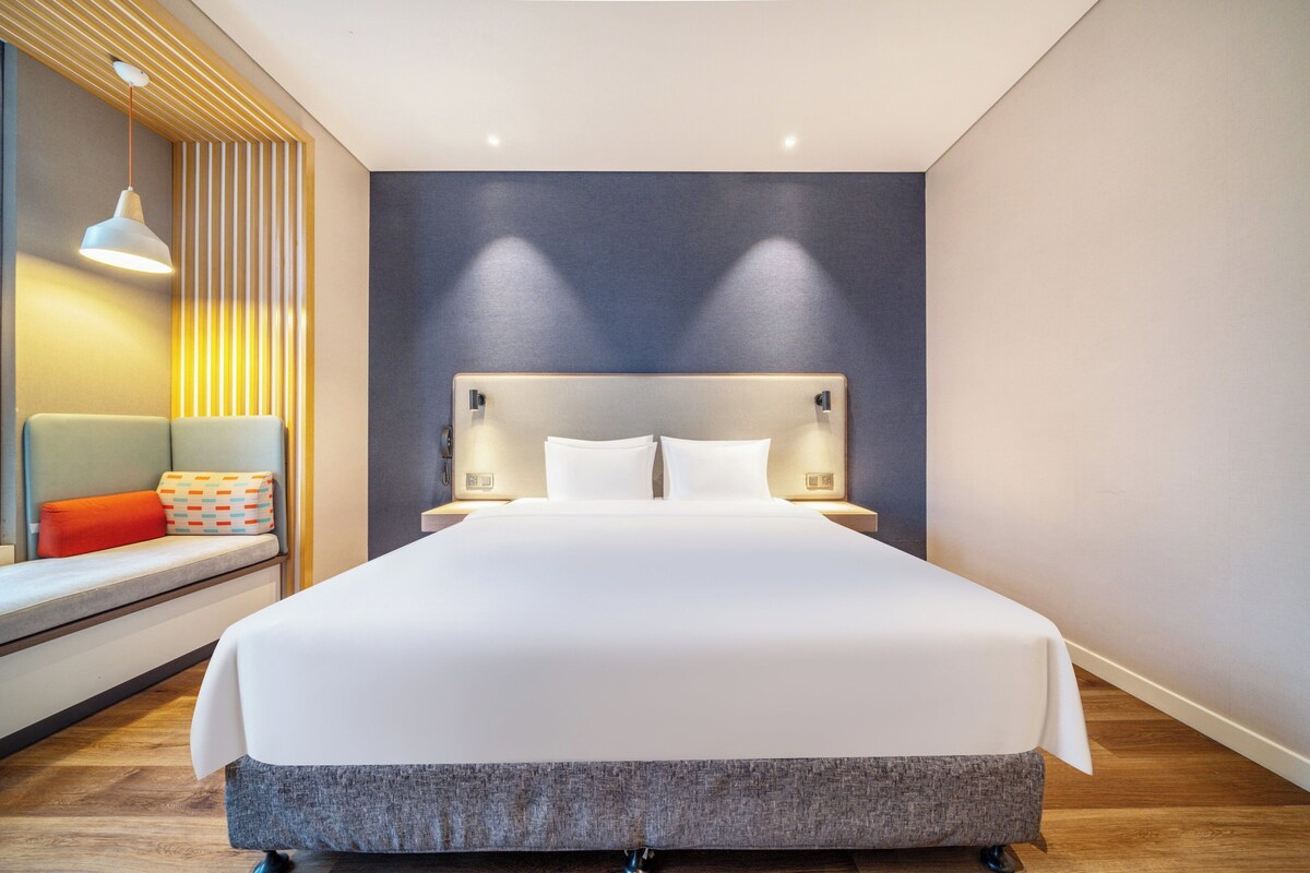 A modern hotel room with a large bed, bedside tables, a bench with cushions, and a wall-mounted light fixture.