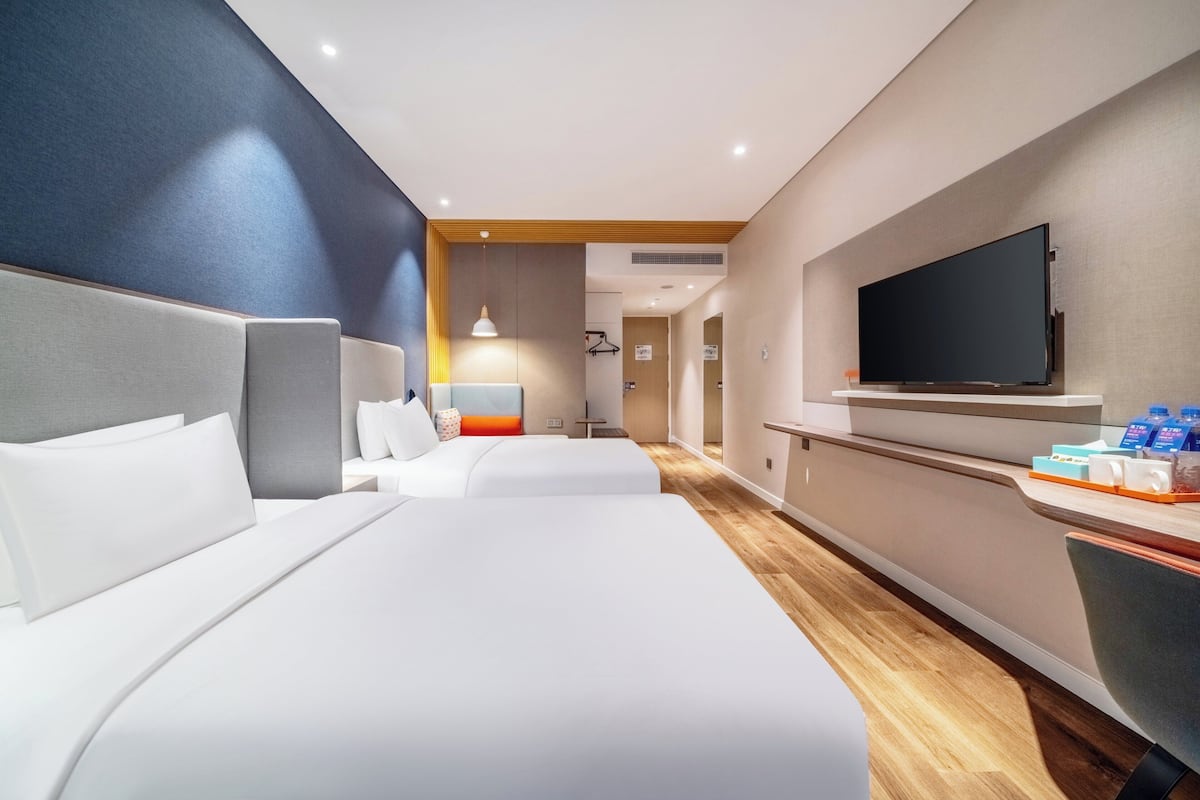 A hotel room with two beds, a TV, a desk with amenities, and a hallway leading to other rooms.