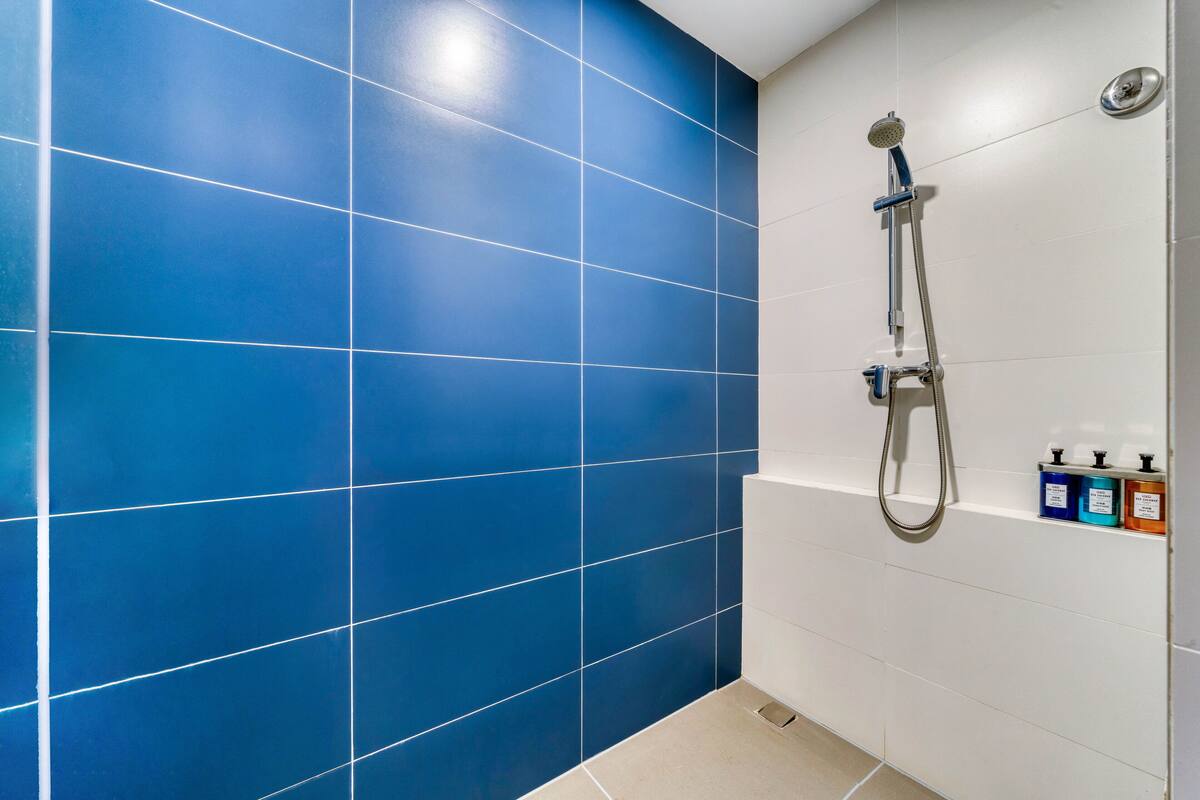 A bathroom with blue tiles, a shower, and a shelf with toiletries.