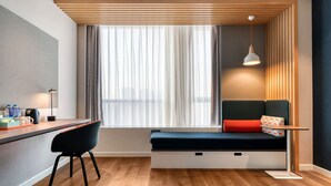 A modern room with a wooden floor, a desk with a chair, a bench with cushions, a hanging light, and a window with curtains.