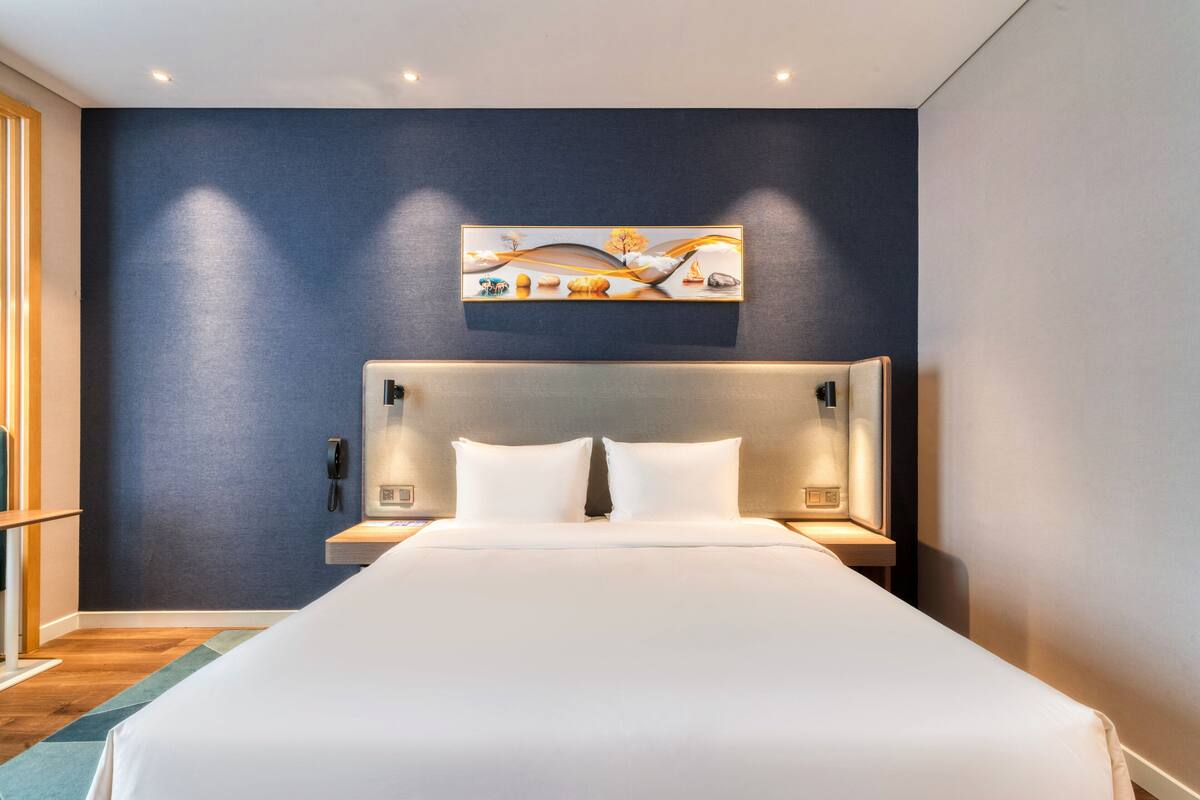 A modern hotel room with a large bed, a headboard with integrated lighting, a bedside table with a phone, and a wall-mounted artwork.