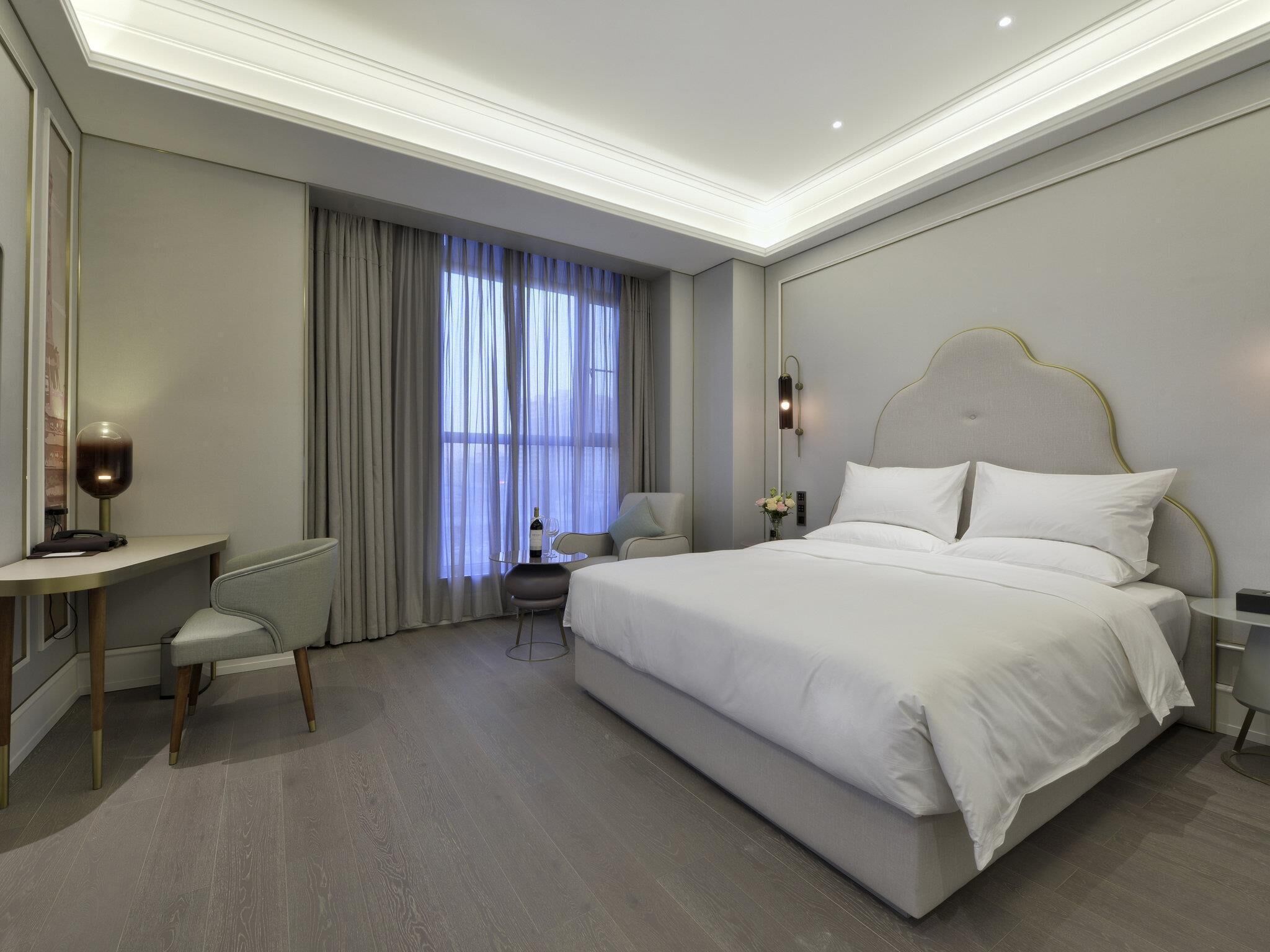 Moderni hotellihuone, jossa on suuri sänky, työpöytä, tuoli ja ikkunasta avautuva näkymä kaupunkiin.