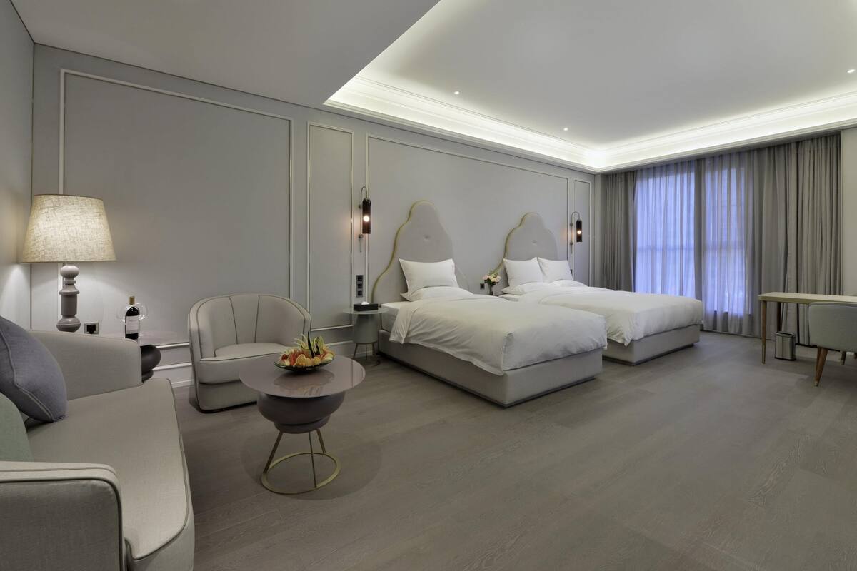 Une chambre d’hôtel moderne avec deux lits, un canapé, une petite table ornée d’un vase de fleurs et une lampe.