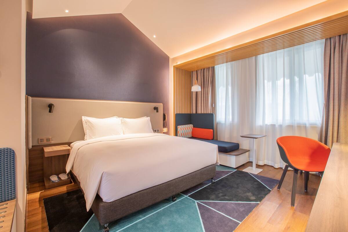 Ein modernes Hotelzimmer mit einem großen Bett, einer kleinen Sitzecke und einem hellen Fenster mit Vorhängen.