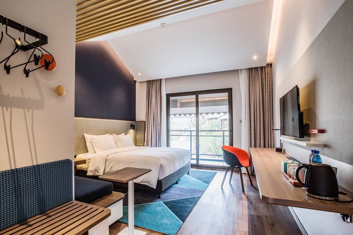 Ein modernes Hotelzimmer mit Bett, Schreibtisch, Stuhl, Fernseher und einem Fenster mit Vorhängen.