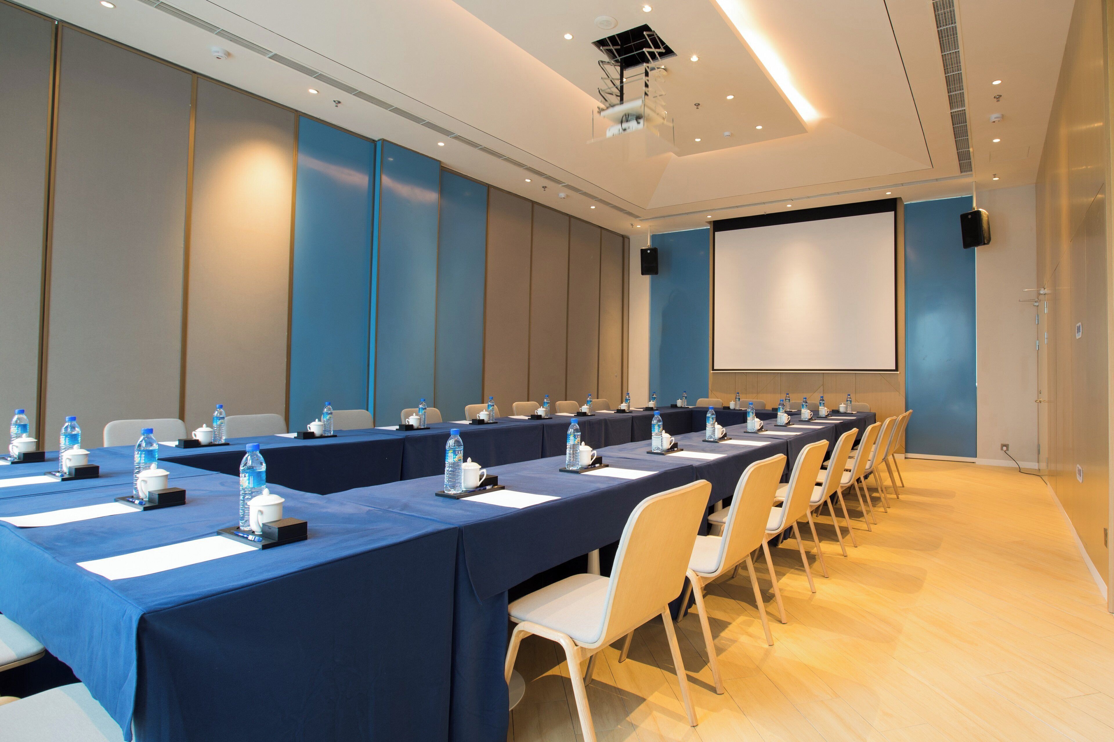 Un salón de conferencias con largas mesas, sillas, botellas de agua y una pantalla de proyector.