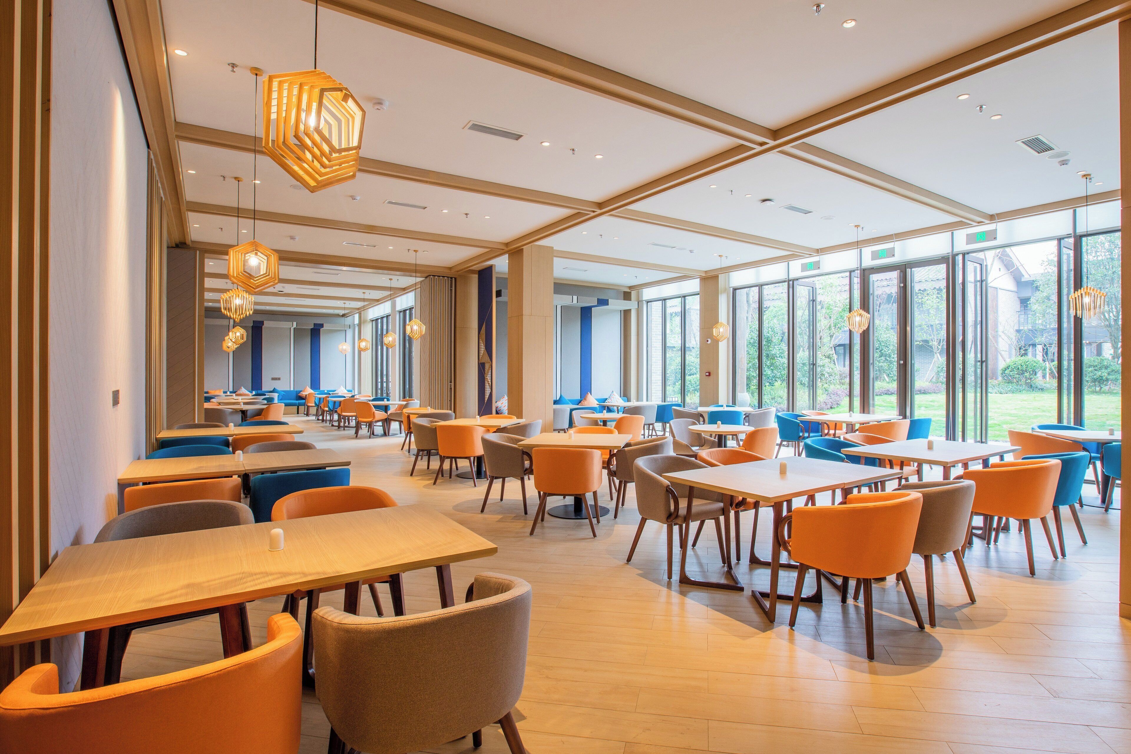 A spacious indoor area with multiple tables and chairs, large windows, and modern lighting fixtures.