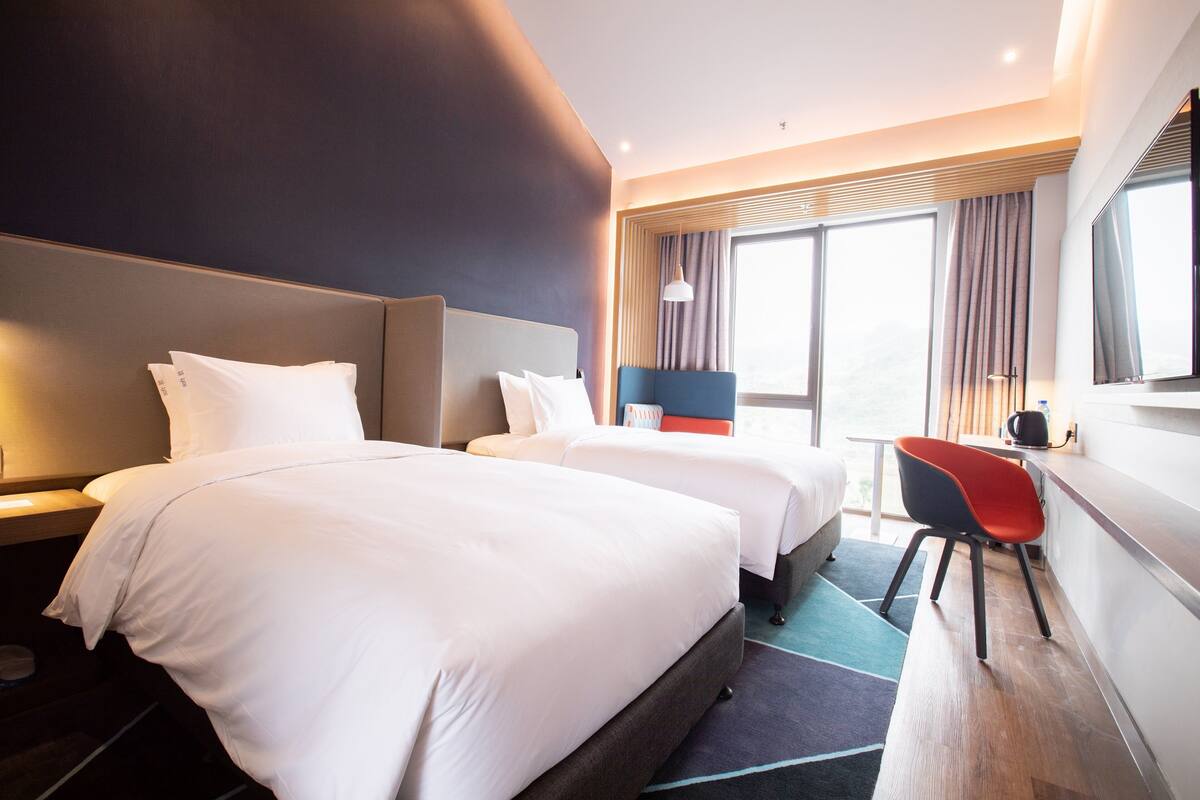 Ein Hotelzimmer mit zwei Betten, einem Schreibtisch, einem Stuhl und einem großen Fenster.