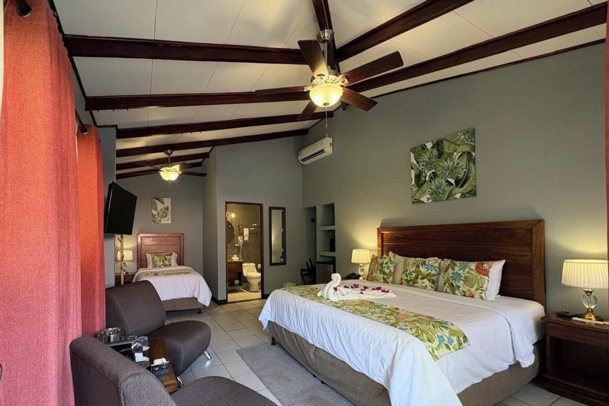 A hotel room with a bed, two chairs, a TV, a ceiling fan, and an air conditioning unit.