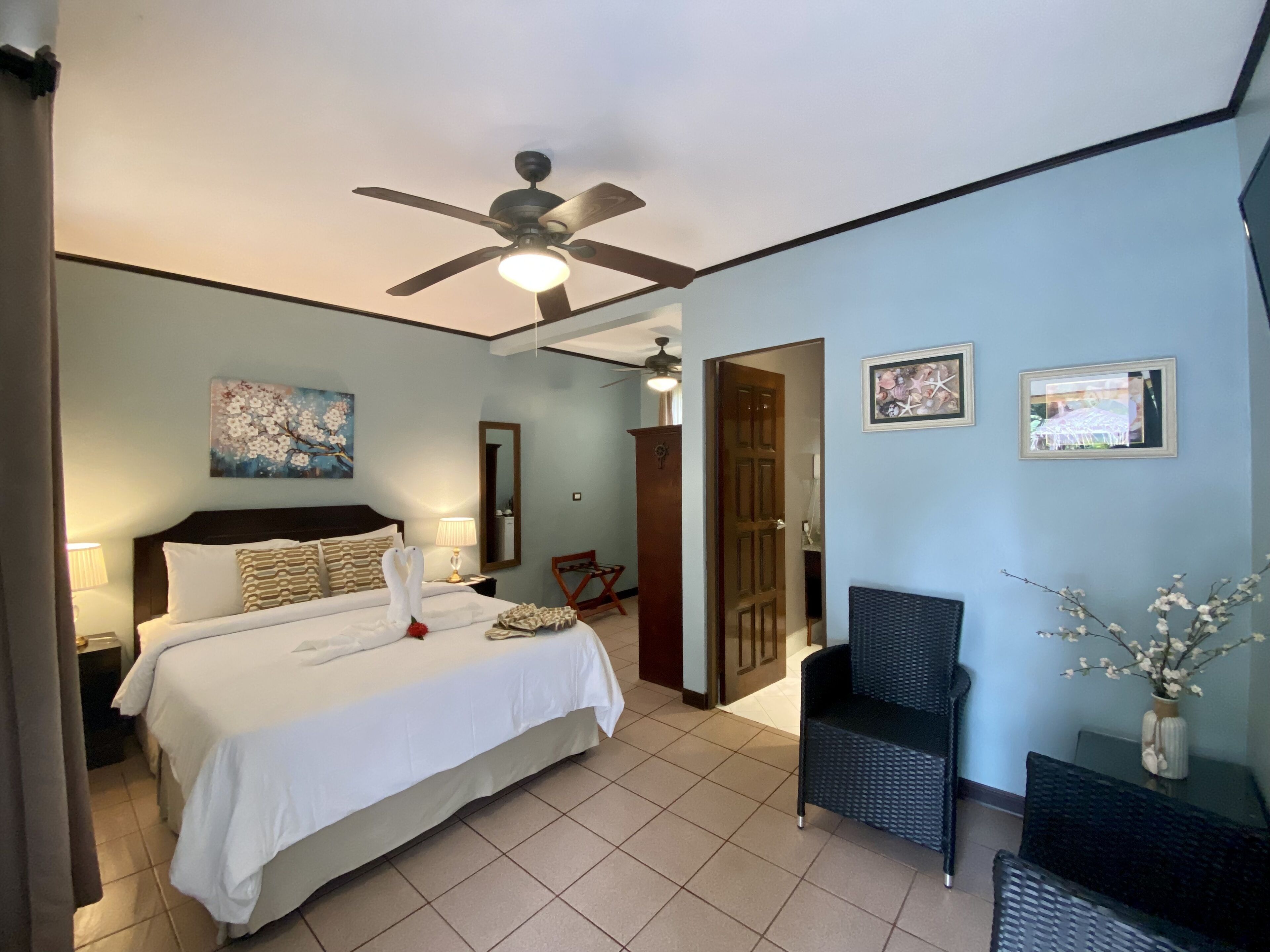 Ein Hotelzimmer mit einem Bett, einem Deckenventilator, einem Nachttisch, einem Sessel und einer Tür zu einem anderen Raum.