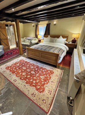Un dormitorio con cama de madera, alfombra estampada en tonos rojo y beige, mesita de noche de madera y una ventana con cortinas.