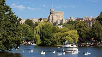 A castle with a round tower, surrounded by lush greenery and a calm river with swans and a boat.