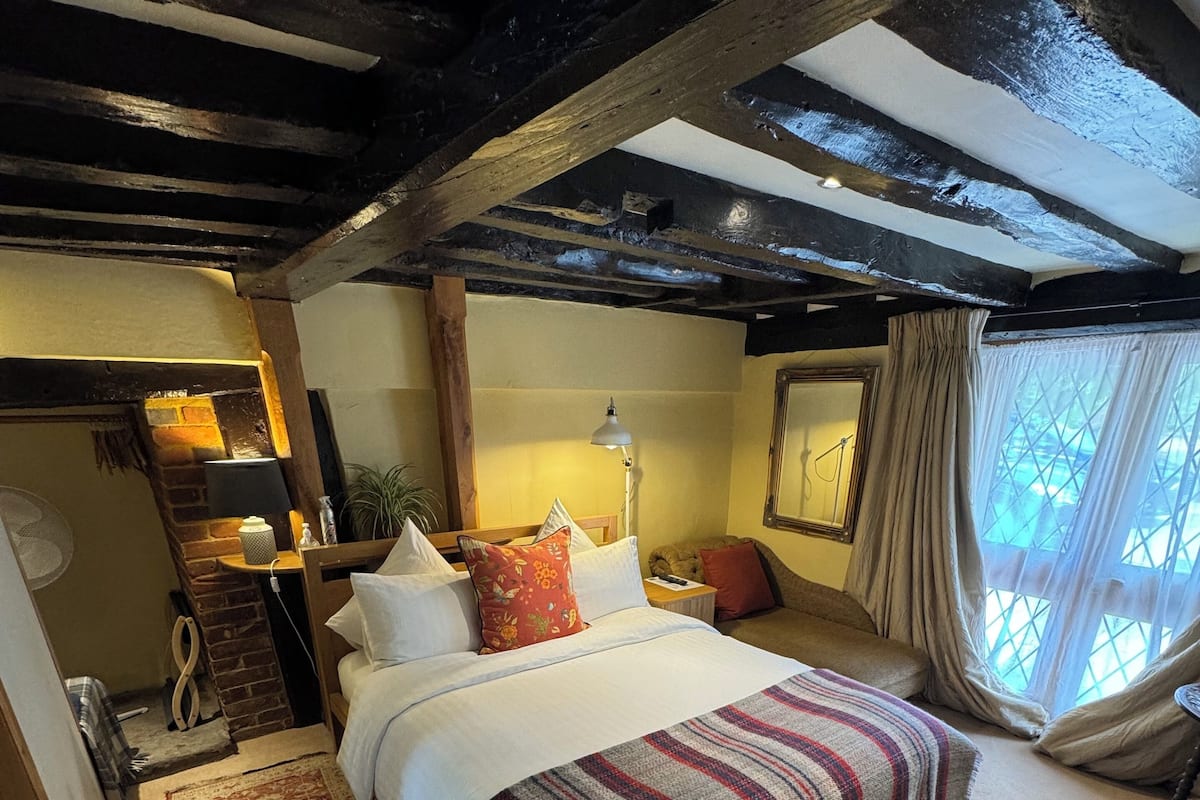 A cozy bedroom with a large bed, a wooden headboard, a nightstand, a lamp, and a window with curtains.