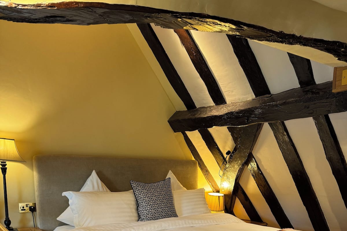 A bedroom with a bed, bedside lamps, a wooden bench, and exposed wooden beams.