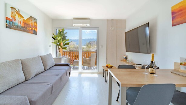 Una sala moderna con un sofá, una mesa de comedor con sillas, un televisor montado en la pared y un balcón con vistas a las montañas.