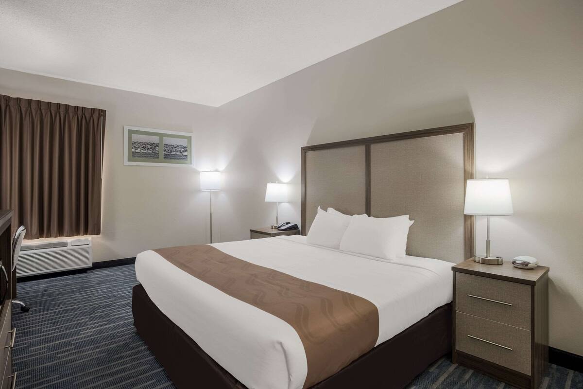 Habitación de hotel con una cama grande, dos mesitas de noche con lámparas, un escritorio y una ventana con cortinas.