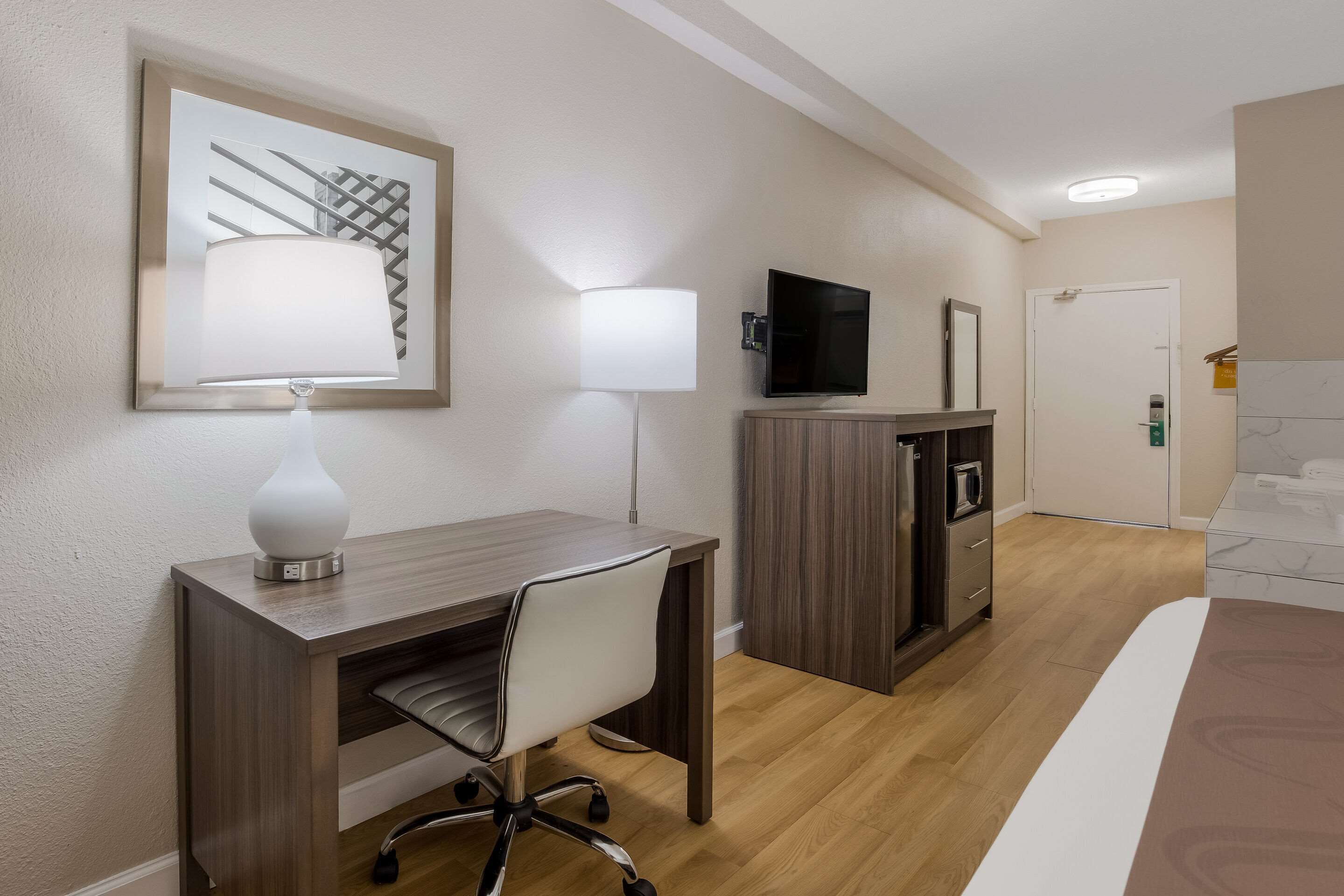 A hotel room with a wooden desk, a chair, a television, a minibar, and a bed.